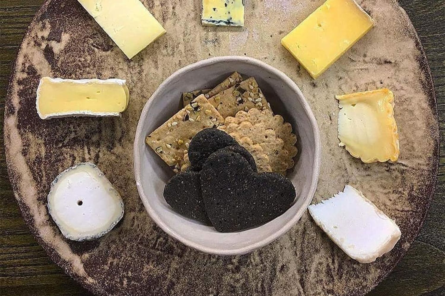 A circular cheese plate with black and brown crackers in a central white dish.