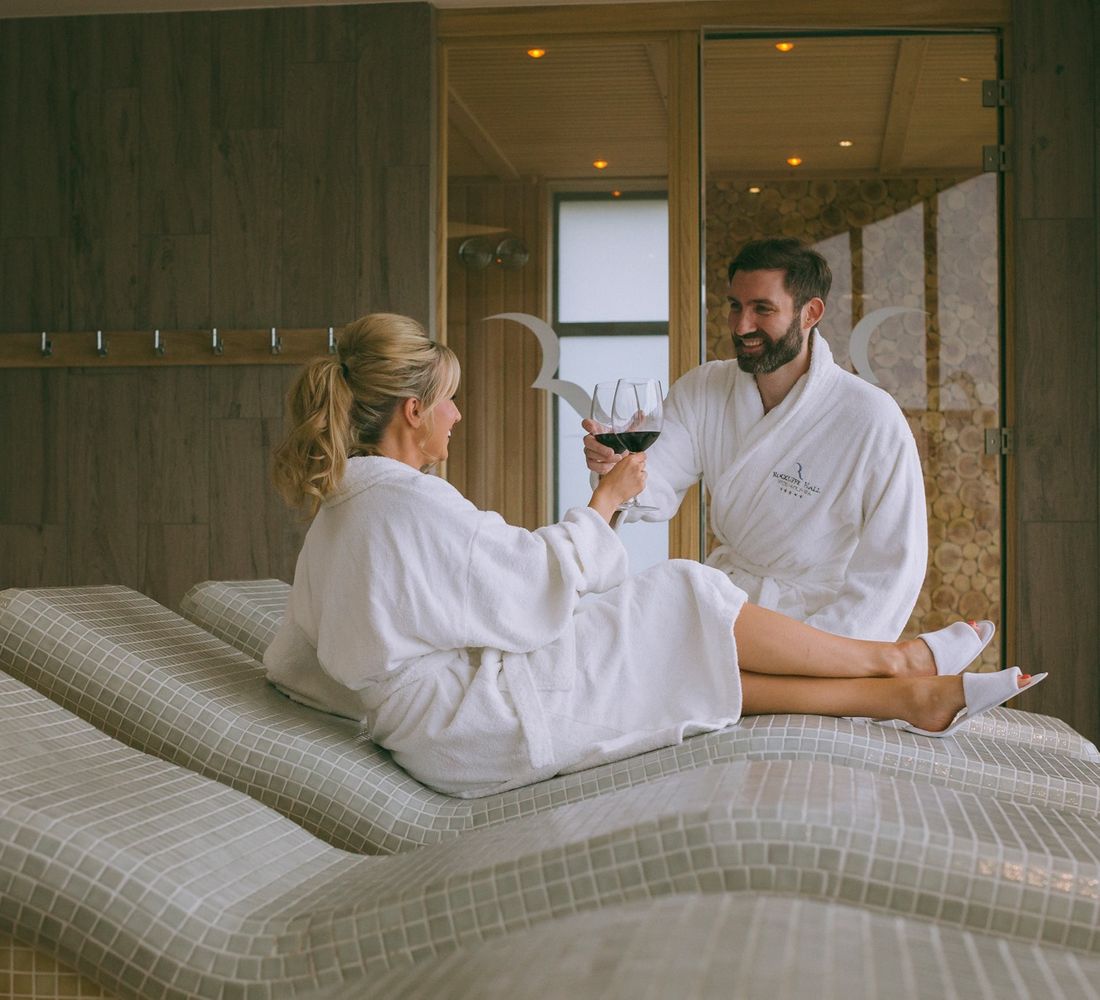 A couple give cheers with red wine while reclined in white robes on Rockliffe Hall spa beds.