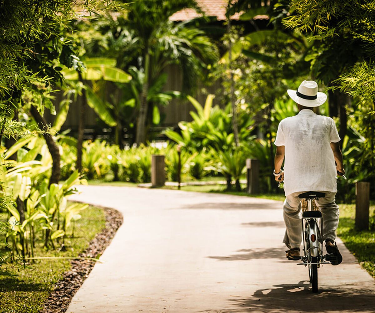A man in a white shirt and with a hat cycles along a wide path. Cambodian plant life and native trees line the sides of the path.