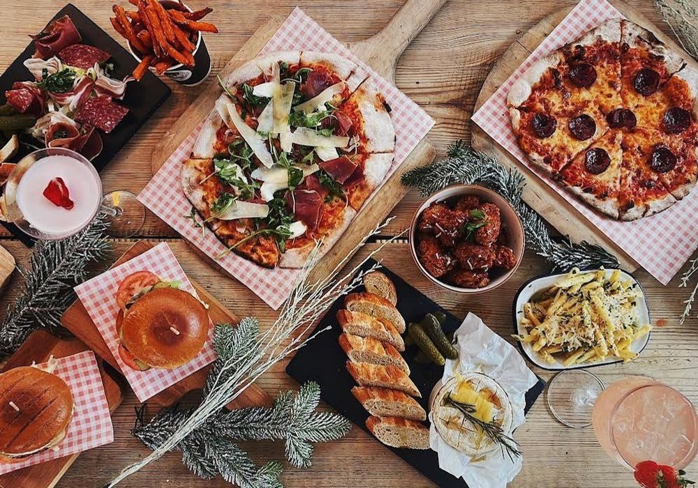 Neverland’s Winterland has a varied food menu to taste. This image shows an aerial view of a selection of dishes - including woodfire cooked pizza on a wooden serving platter and white and red checked paper.