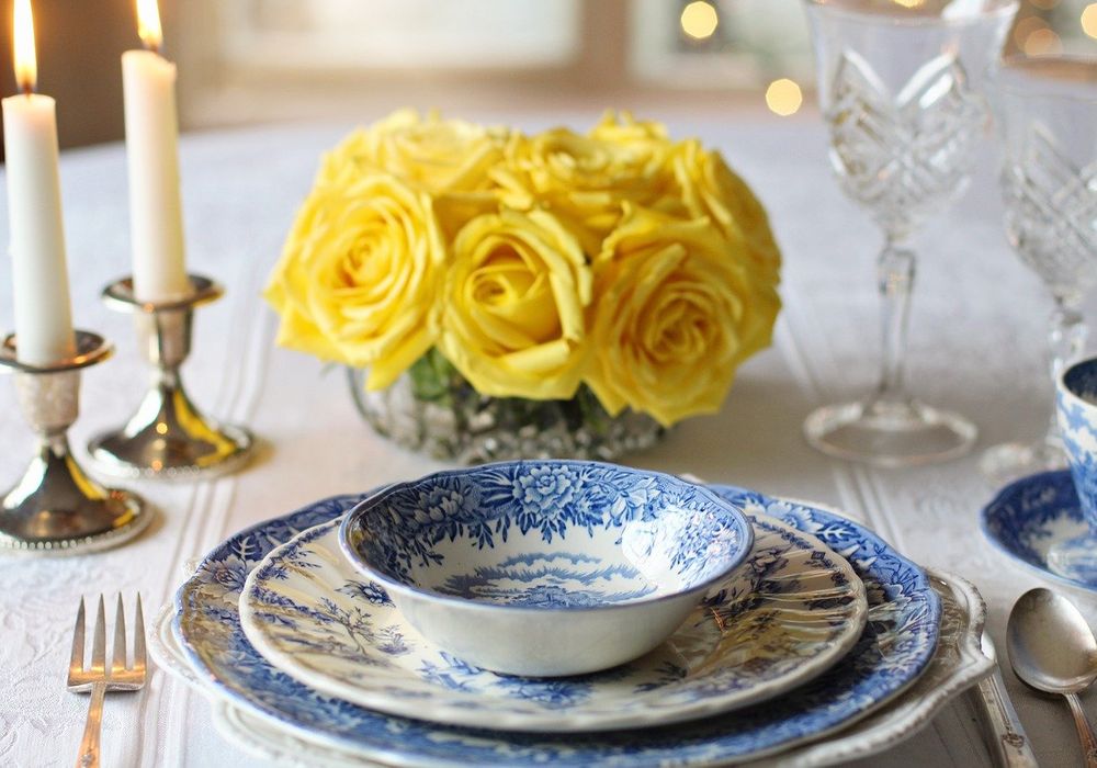 A blue and white dining set sits on a white table cloth with yellow flowers and candles arranged.